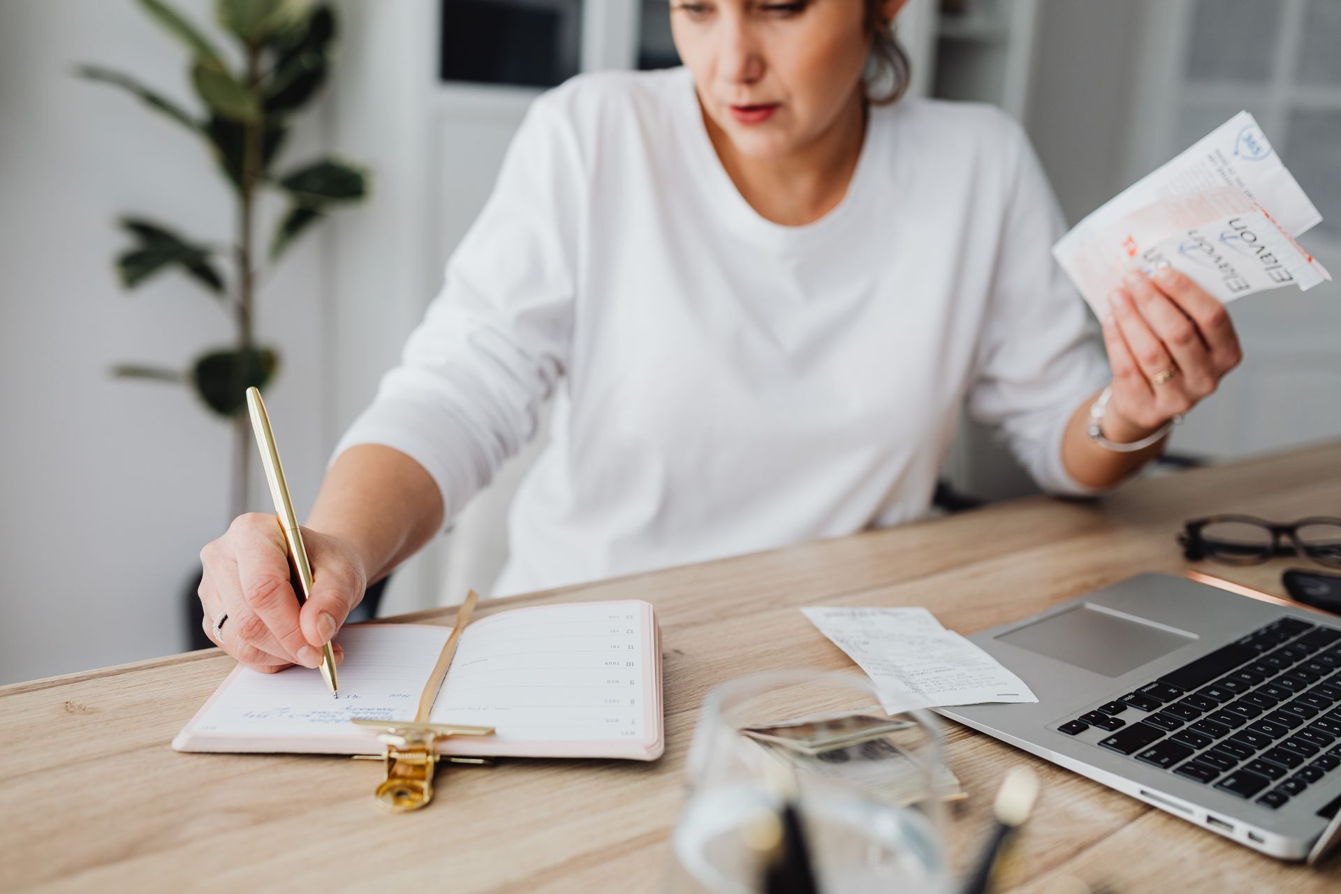 Self employed woman recording expenses in a notebook beside receipts and a laptop