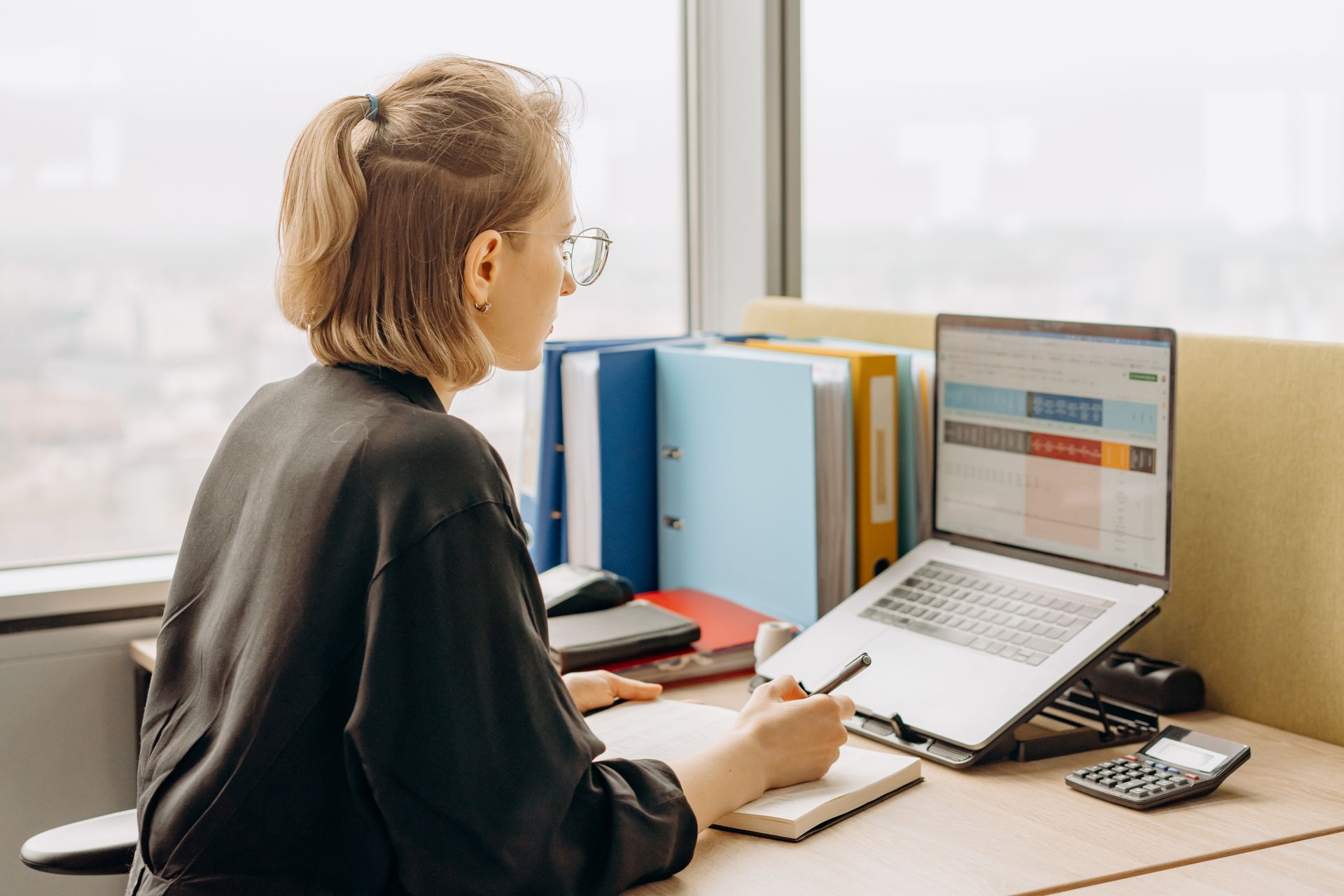 Woman working on online bookkeeping services at a laptop, with binders and a calculator on the desk