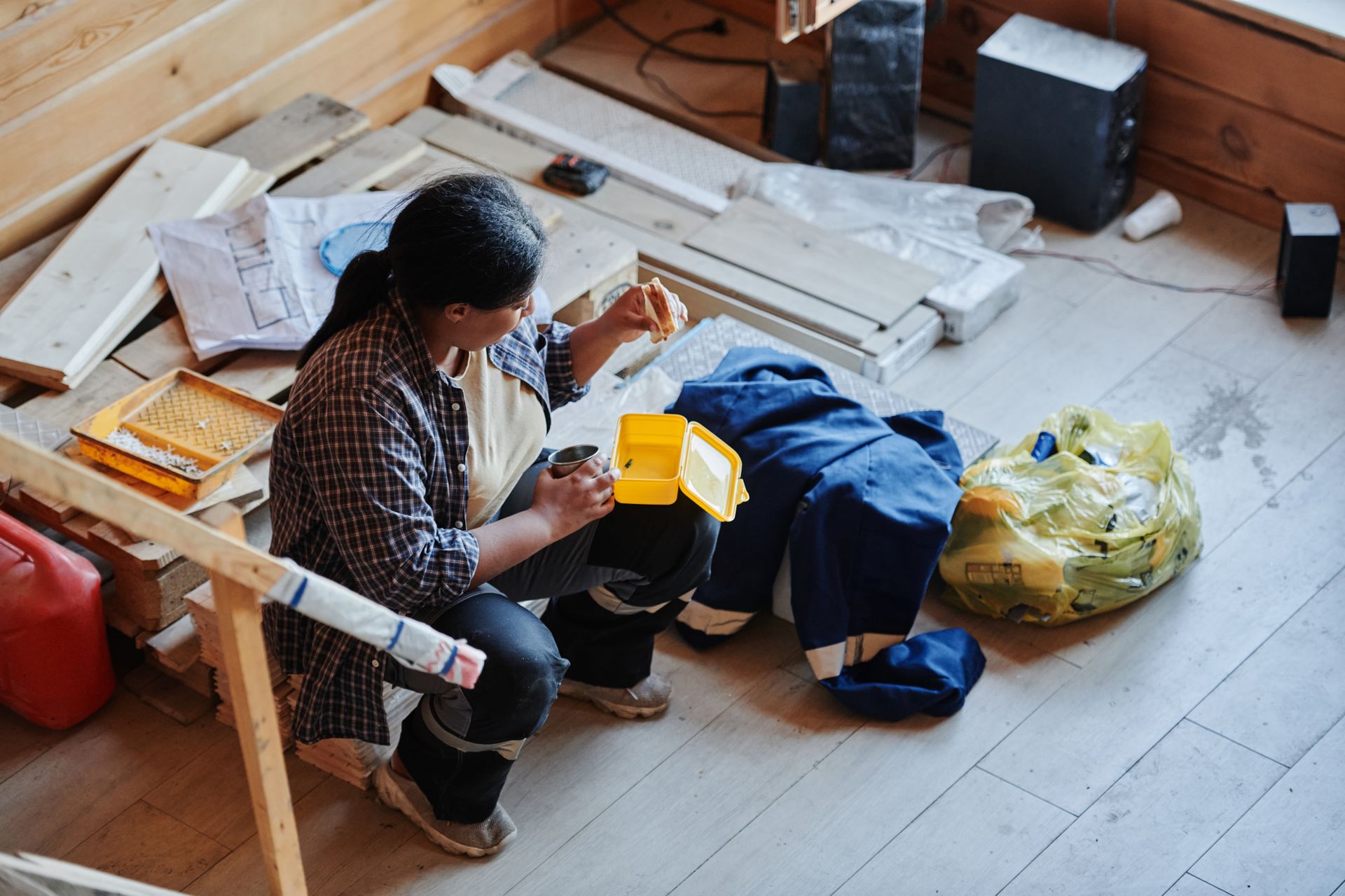 Worker in a plaid shirt eating a sandwich from a yellow lunch box among lumber on a construction site